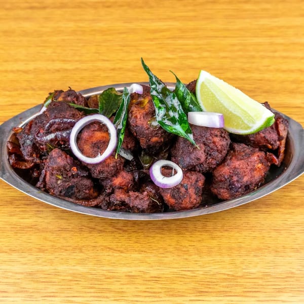 Spicy chicken 65, garnished with curry leaves, red onion rings, and a lime wedge, on a metal platter.