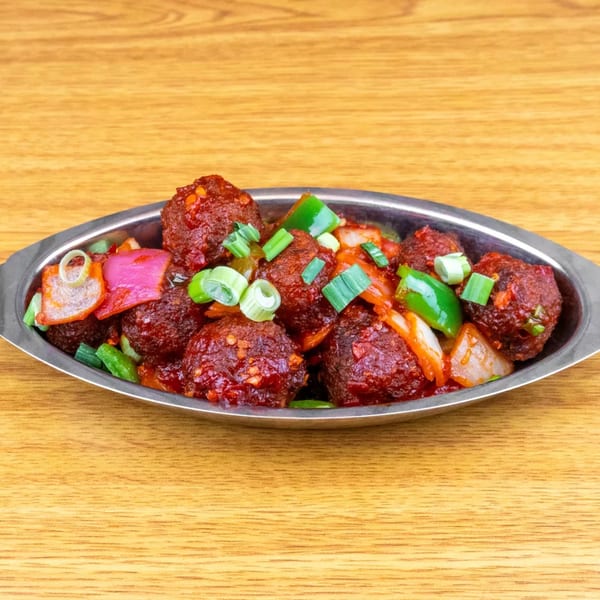 Manchurian dish in a silver bowl, with red sauce, green onions, and bell peppers on a wooden table.