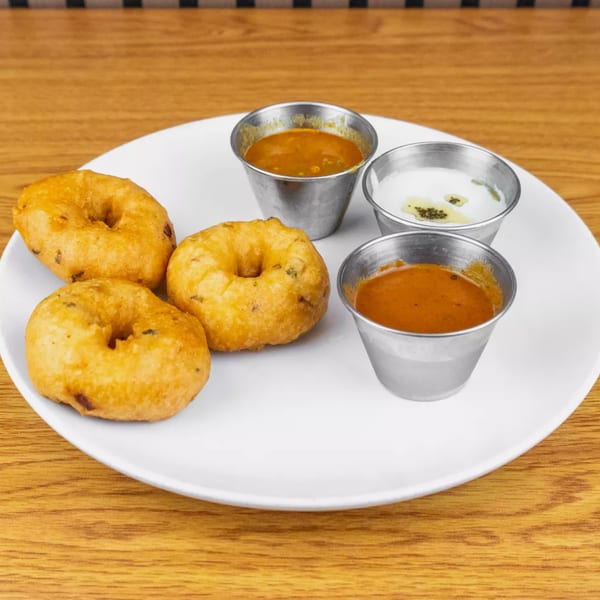 Three golden-brown vada donuts with three small metal cups of dipping sauces on a white plate.
